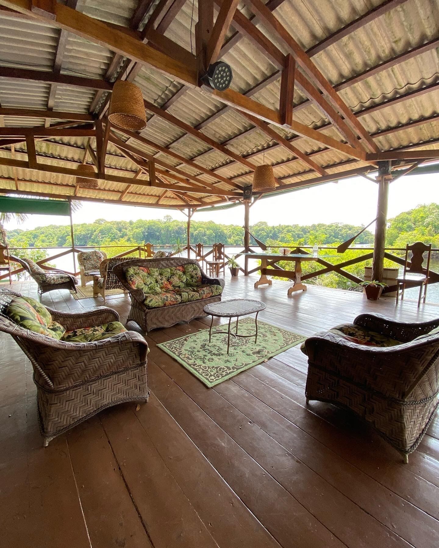 Uma área de estar coberta ao ar livre com móveis de vime e vista para uma paisagem arborizada.