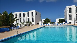 Una piscina con baldosas azules, rodeada de sillones de descanso y palmeras, con edificios blancos al fondo.