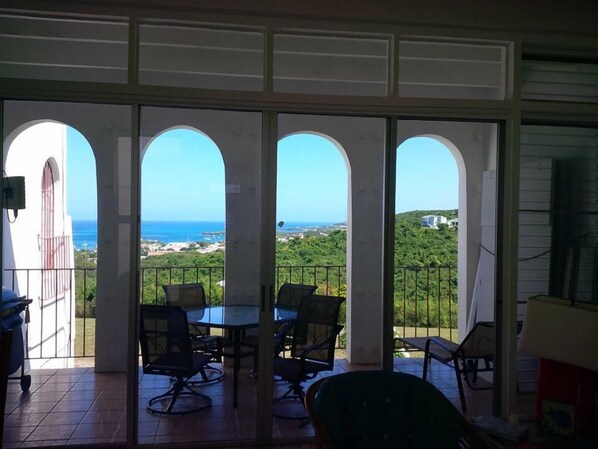 A balcony with a view of the sea and greenery. There are four chairs and a table.