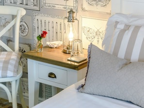 Una cama bien tendida con un almohadón a rayas, una mesita de noche con una lámpara, un florero con una rosa y una pared con diseños decorativos.
