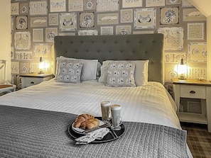 Una cama bien hecha con almohadones estampados, una bandeja con cruasanes y café, y una pared decorada con cuadros.