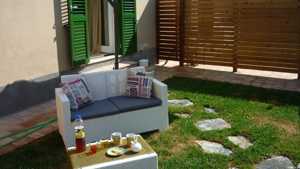 A patio with a white wicker sofa, a table with drinks and a teapot, and a white umbrella providing shade.