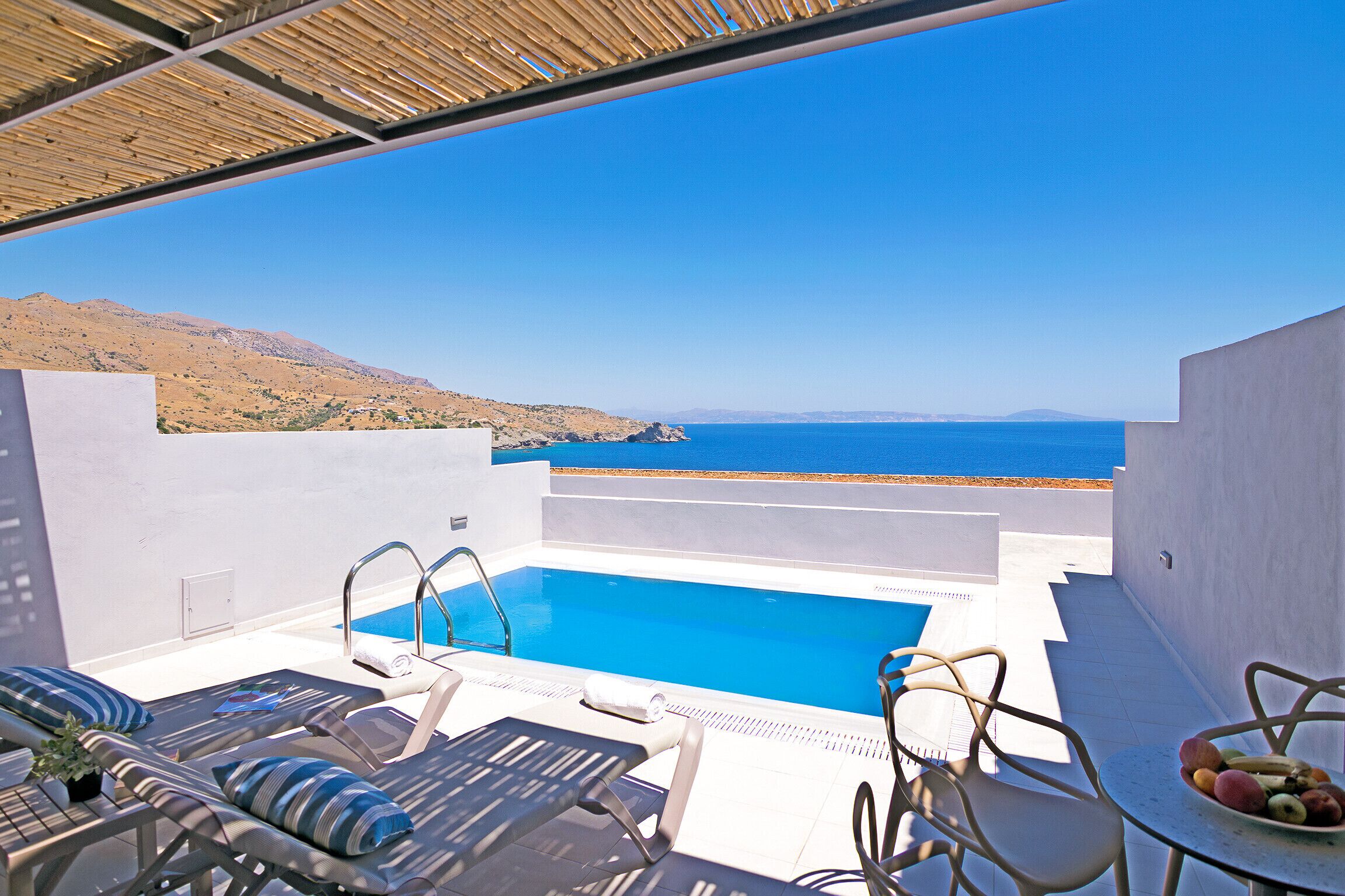 Área na piscina com vista para o mar, cadeiras de espreguiçadeira e uma mesa com frutas.