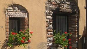 Un edificio con due finestre ad arco, una con facciata in pietra e l’altra in mattoni. Entrambe le finestre presentano balconiere con fiori rossi.