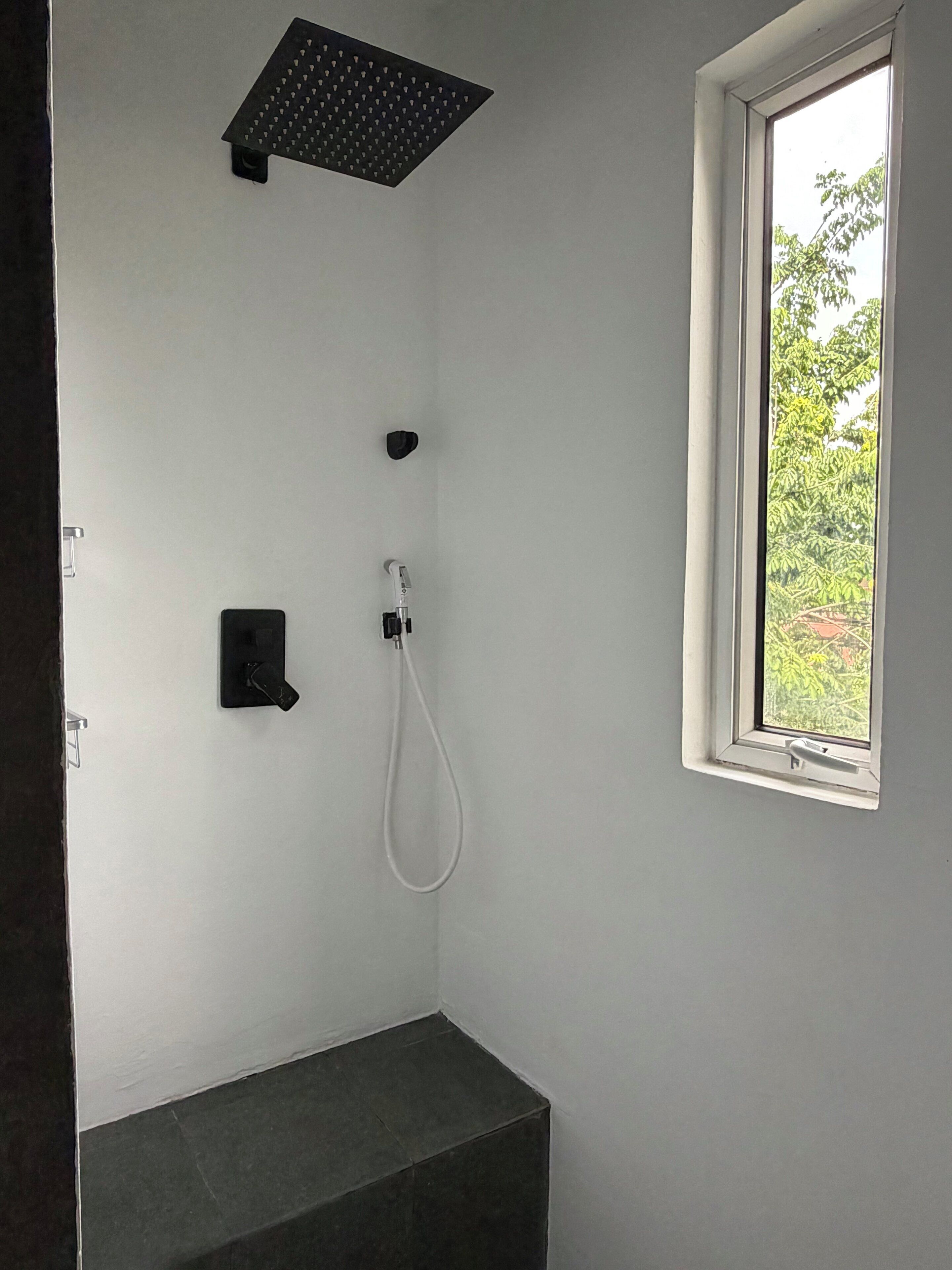Een douche met een handdouche, een bedieningspaneel aan de muur en een raam met uitzicht op groen.