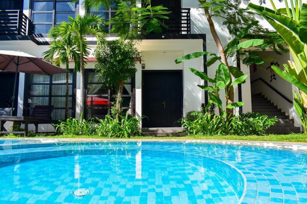 Una piscina con bordes de azulejos, rodeada de exuberante vegetación y un edificio moderno con una escalera y una puerta.