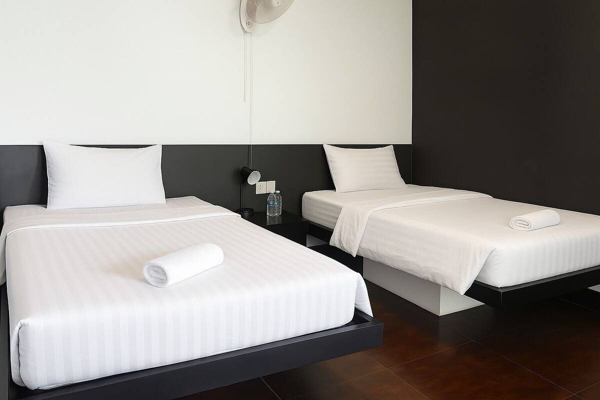 A hotel room with two beds, each with white linens and pillows, a nightstand with a water bottle, and a wall-mounted light fixture.