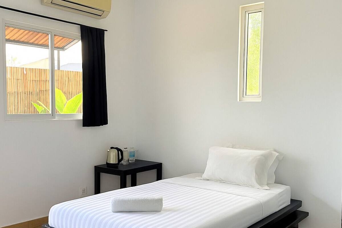 A single bed with a white mattress and a dark wooden frame, a ceiling fan, an air conditioning unit, a small table with a kettle, and a window with a view of greenery.
