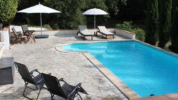 A swimming pool with lounge chairs and umbrellas, surrounded by stone flooring and greenery.