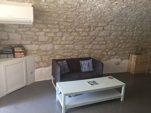 A living room with a stone wall, a dark brown sofa with patterned cushions, a white coffee table, and a small wooden cabinet. - Traditional 1 bed French gite, with private pool, 3 acres of garden. Aircon. (Valprionde)