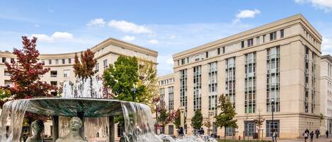 Una fuente pública con juegos de agua y estatuas, rodeada por una plaza con edificios al fondo.