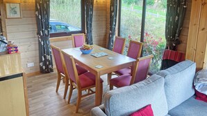 A wooden cabin interior with a dining table, chairs, a sofa, and large windows with curtains.