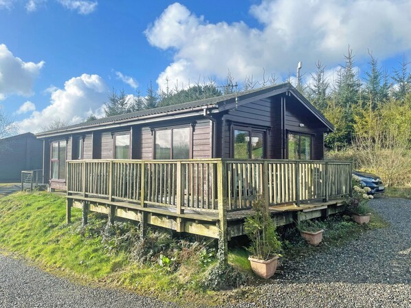 A wooden cabin with a deck, large windows, and a gravel driveway.