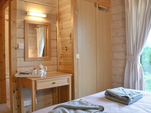 A wooden room with a bed, a bedside table with a lamp, a mirror, and a window with curtains.