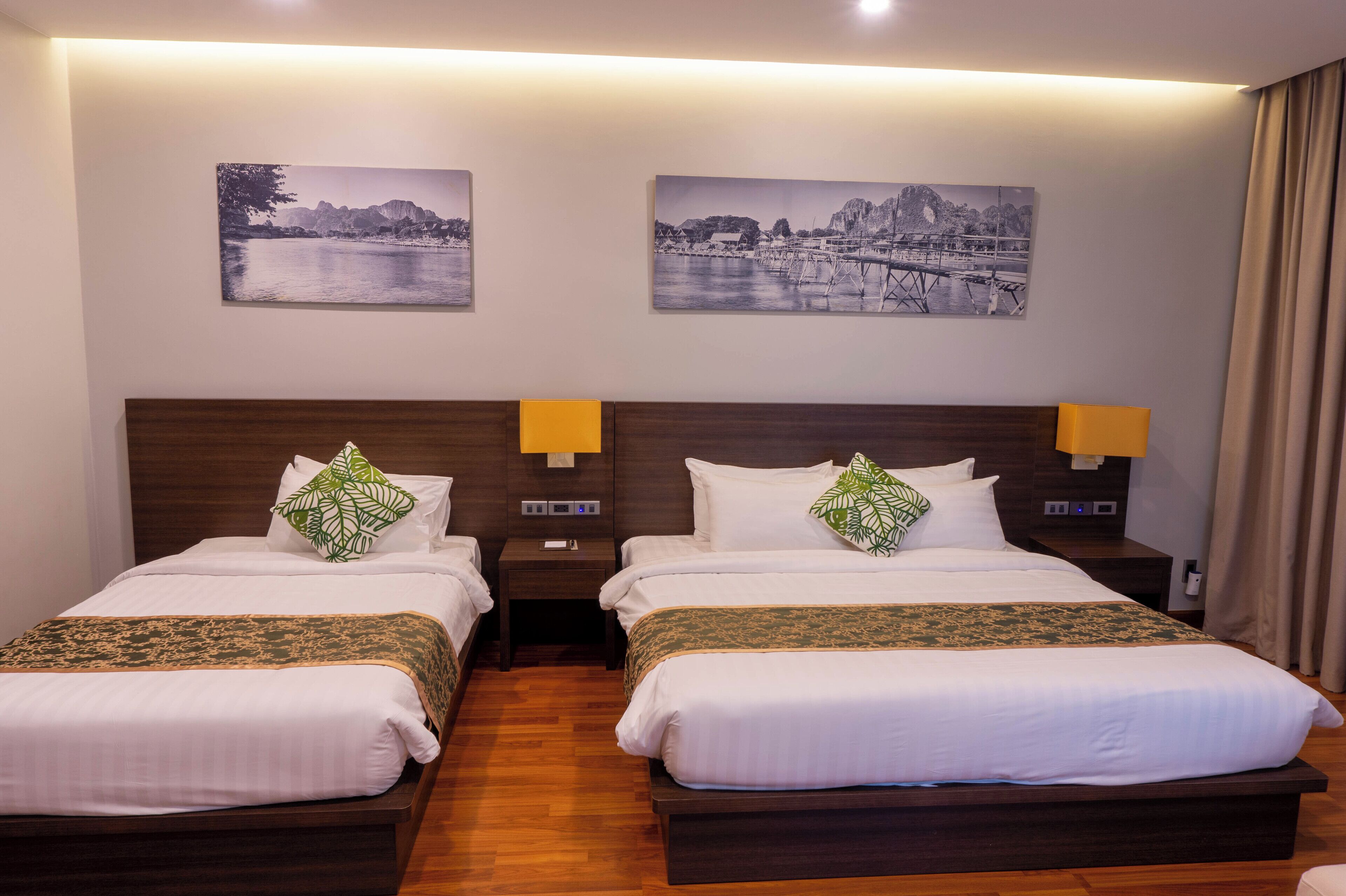 A hotel room with two beds, wooden headboards, white bedding, green and gold throw pillows, nightstands with lamps, and two black-and-white landscape paintings on the wall.