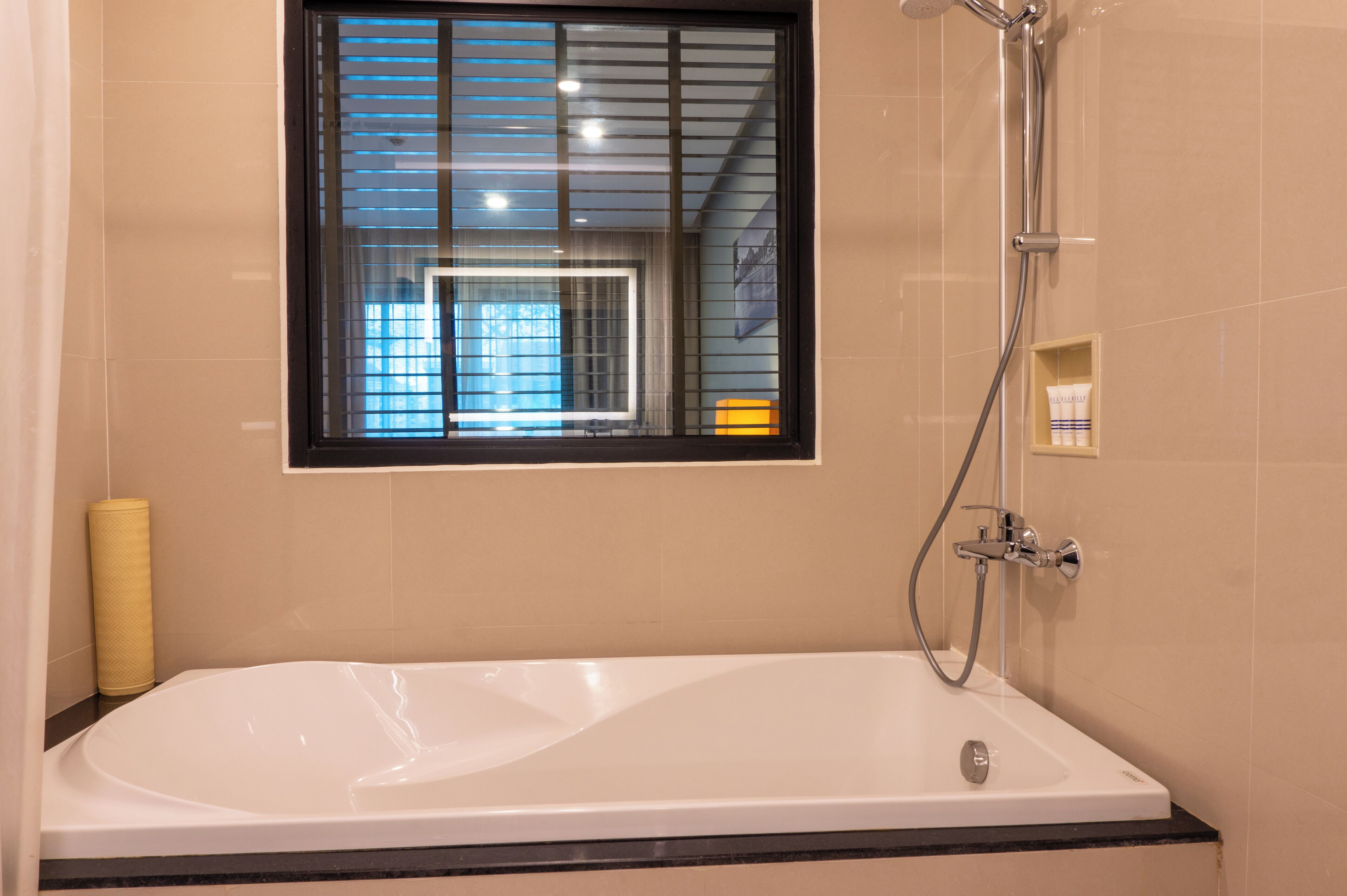 A modern bathroom with a bathtub, a window with blinds, a showerhead, and a small shelf with toiletries.