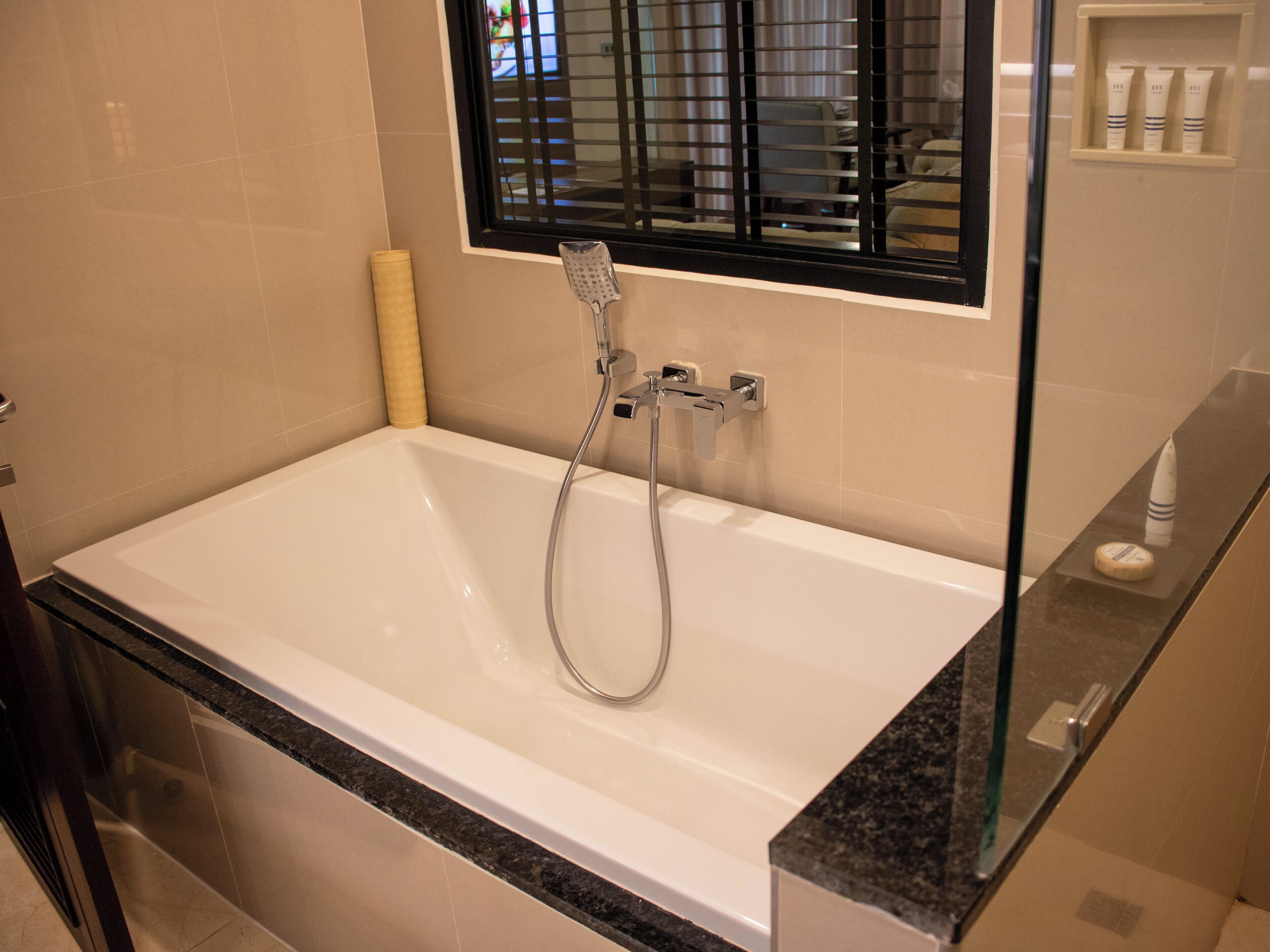 A modern bathroom with a large bathtub, a showerhead, and a window with blinds.
