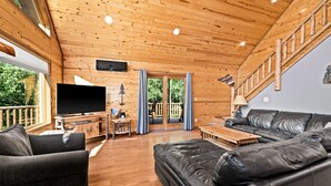 A wooden cabin interior with a TV, leather sofas, a wooden table, and a balcony with a view of greenery.