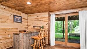 A wooden interior with a rustic bar area, wooden chairs, and a view of greenery outside.