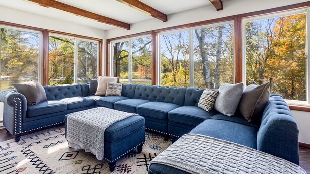 A large, L-shaped sectional sofa with multiple cushions and a matching ottoman, positioned in a room with large windows offering a view of trees and a clear sky.