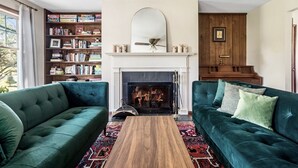 A living room with two green sofas, a wooden coffee table, a fireplace, a bookshelf, and a piano.