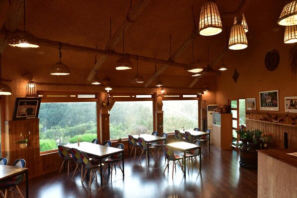 A restaurant with wooden tables and chairs, large windows with a view of greenery, and hanging pendant lights. - Golmangtae (Boseong)