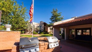 A barbecue grill setup with a flag, brick wall, and a building with a tiled roof in the background. - Johnny's Sedona Getaway - 2 Primary Suites 2 Bath (Sedona)