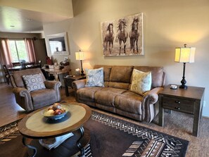 Un salon avec un canapé en cuir marron, deux fauteuils, une table basse sur laquelle on a disposé des fruits, un tapis et un tableau représentant des chevaux accroché au mur.