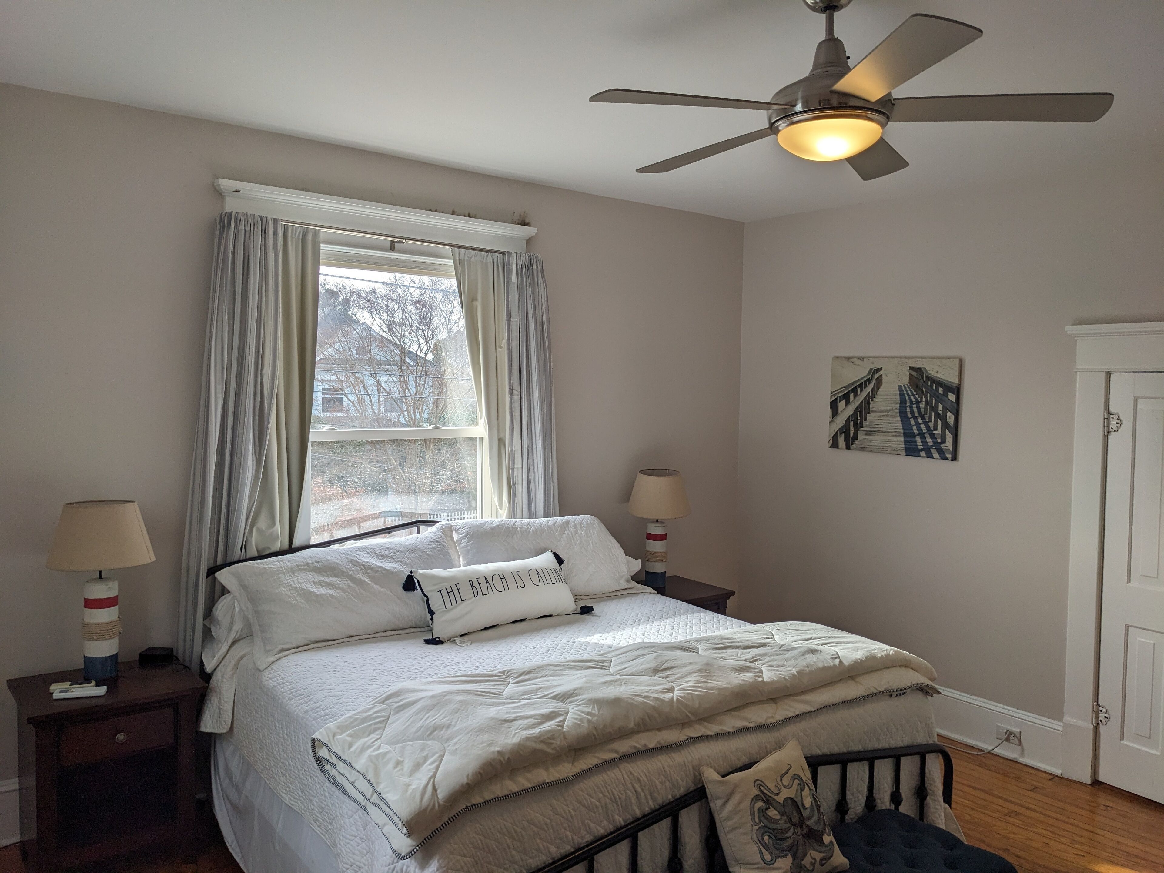 Un dormitorio con cama, ventilador de techo, ventana con cortinas, lámparas de noche y un cuadro colgado en la pared.