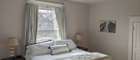 Un dormitorio con cama, ventilador de techo, ventana con cortinas, lámparas de noche y un cuadro colgado en la pared.
