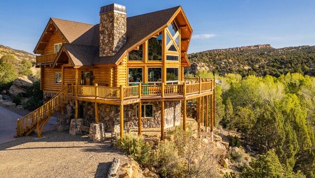 A log cabin with a stone chimney, multiple decks, and a scenic mountain backdrop. - Jewel of Escalante LLC (Escalante)