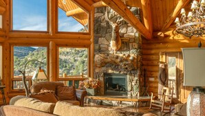 A log cabin interior with a stone fireplace, wooden beams, and a view of the mountains. - Jewel of Escalante LLC (Escalante)