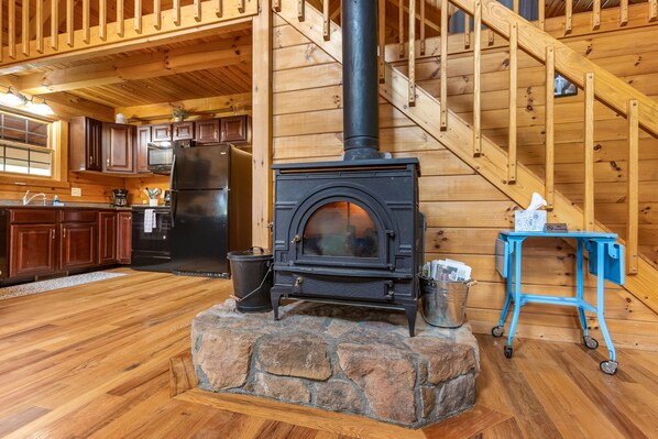 Interior rústico de una cabaña con chimenea de piedra, suelo de madera y una zona de cocina con armarios y electrodomésticos de madera.