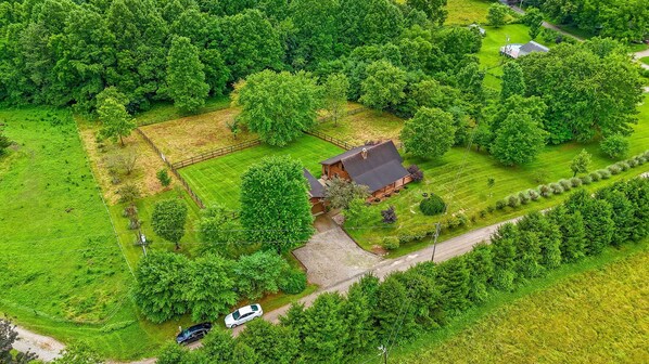 An aerial view of a rural property with a large house surrounded by lush greenery, a fenced yard, and a gravel driveway.