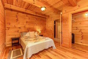 A wooden cabin bedroom with a bed, nightstands, a lamp, and a door leading to another room. - White Pine - Cozy Cabin with Relaxing Hot Tub! (Nelsonville)