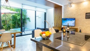 A modern kitchen with a large island, a dining area with a glass door leading to a pool, and a TV mounted on a brick wall. - Villa between the jungle and the Caribbean Sea, Bahia Principe (Akumal)