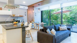 A modern kitchen with a central island, dining area with chairs, a sofa with cushions, and large windows with a view of greenery. - Villa between the jungle and the Caribbean Sea, Bahia Principe (Akumal)