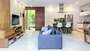 A modern living room with a grey sofa, wooden entertainment unit, and a TV. A kitchen with white cabinets and stainless steel appliances is visible in the background. - Villa between the jungle and the Caribbean Sea, Bahia Principe (Akumal)