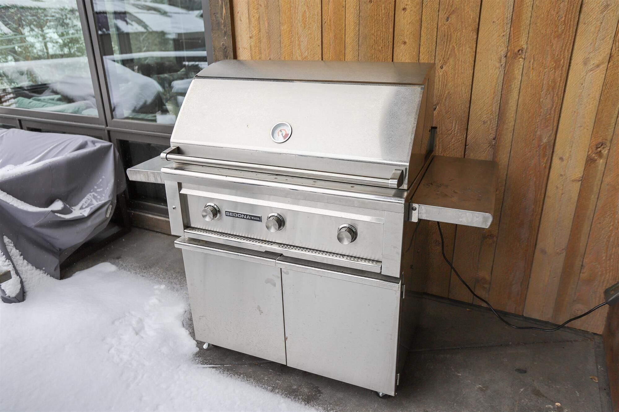 A stainless steel grill with two burners and a side shelf, positioned outdoors against a wooden wall.
