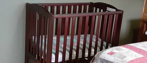 Una cuna de madera con un colchón estampado frente a una pared verde, un suelo de madera y un cuadro enmarcado sobre la cuna.