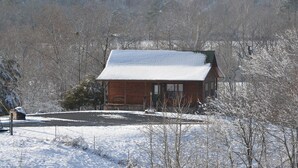 Une cabane en bois, dont le toit est recouvert de neige, entourée d’un paysage enneigé et d’arbres sans feuilles.