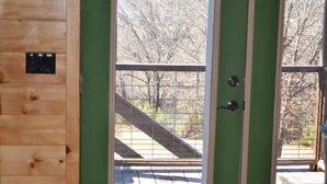 Une porte en bois, dotée d’une poignée en métal et d’un petit panneau de commande, donne accès à un balcon dont le plancher est en bois et la rambarde en métal.
