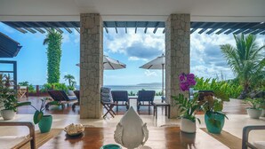 Una mesa de madera con vista al mar, rodeada de sillones de descanso y sombrillas, con plantas en macetas y un pilar de piedra.