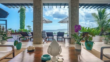 A wooden table with a view of the sea, surrounded by lounge chairs and umbrellas, with potted plants and a stone pillar.