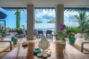 Une table en bois offrant une vue sur la mer, entourée de chaises longues et de parasols, avec des plantes en pot et un pilier en pierre.
