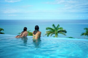 Dos personas en una piscina infinita azul cristalina, con vista al océano y palmeras.