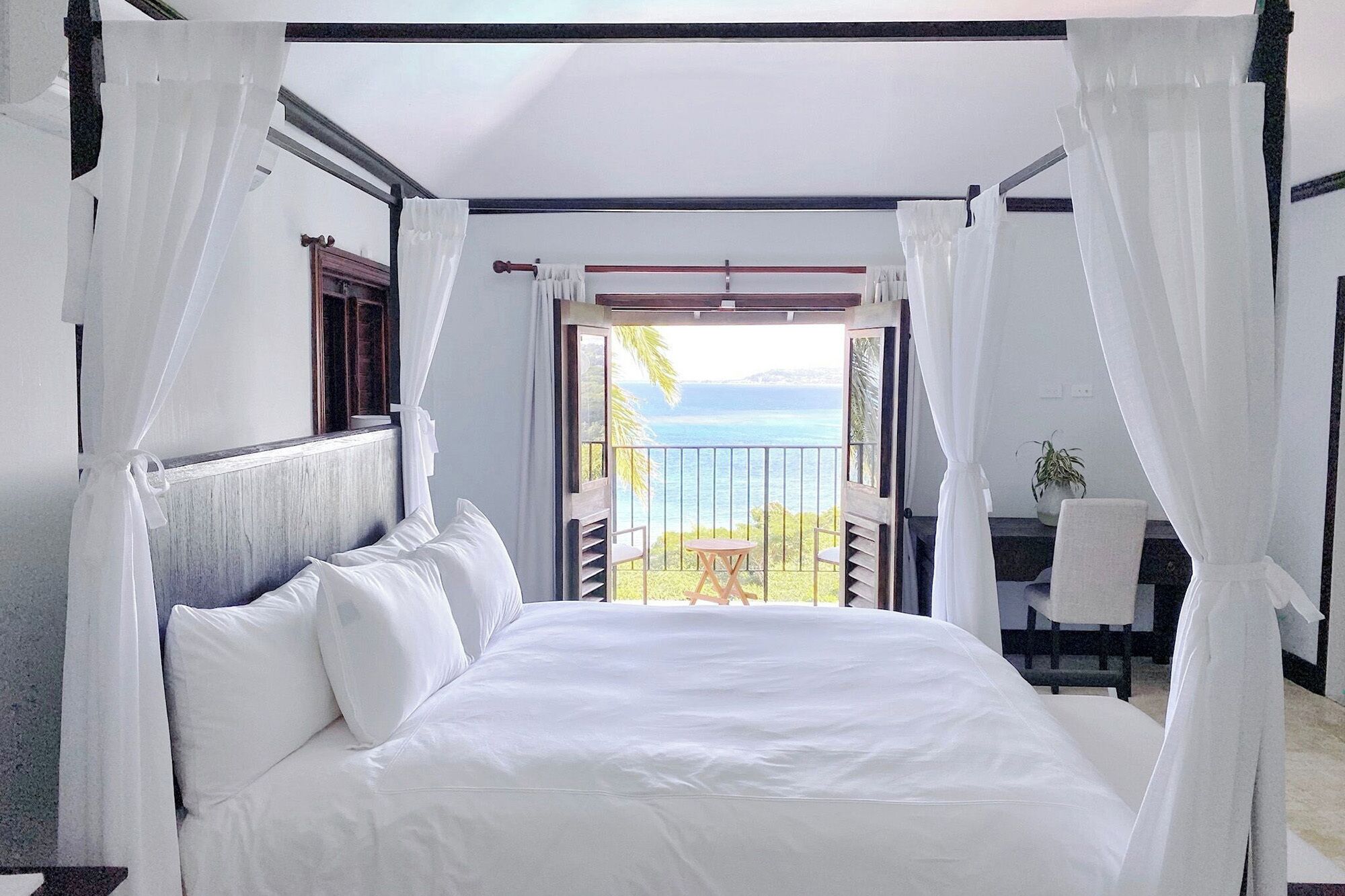 Quarto com cama, cortinas brancas, vista para o mar e varanda com mesa e cadeiras.