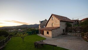 A stone house with a sloped roof, a paved driveway, and a grassy yard with a wooden bench. - Full rental house for 8-10 people, ideal for families with pets. (Ponte-Caldelas)