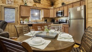 A wooden cabin interior with a dining table set for four, kitchen appliances, and a decorative wall hanging.