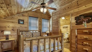 A wooden cabin bedroom with a bed, a dresser, a ceiling fan, and a bathroom visible through an open door.